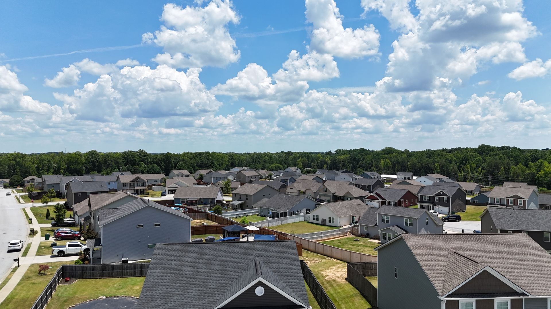 Aerial view of a suburban neighborhood with houses, lawns, and a cloudy blue sky overhead.